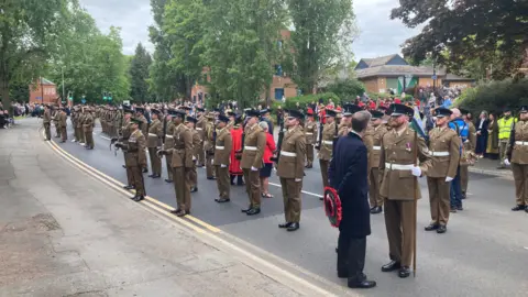 Hundreds of soldiers in brown uniform with white belts stand in line on a road during a town parade.