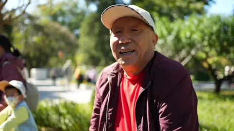 BBC Mr Meng sits in Ritan Park wearing a white hat and lavender jacket.