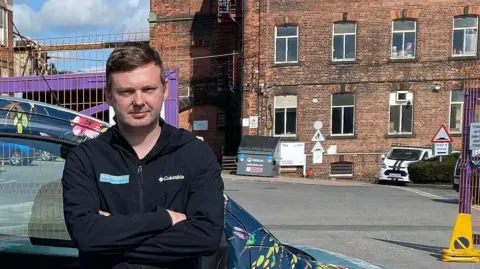 Sam Holey with light brown hair wearing a black fleece top at Fieldhouse Industrial Estate. He has his arms folded and is leaning on his business van. A converted mill is in the background.