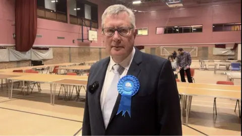 Roger Hirst with grey hair and wearing a navy suit with a white shirt and tie standing an leisure centre hall.