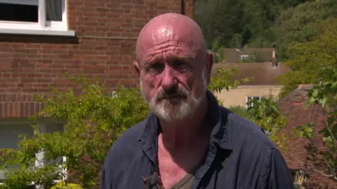 A man with a bald head and a beard in a dark blue shirt. He is stood in front of a building, with a small tree in the background.