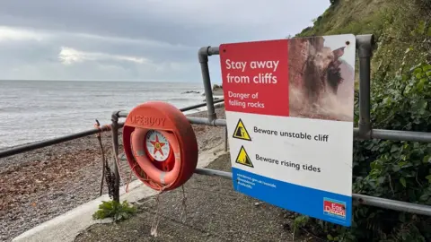 Miles Davis/BBC A sign warning people to stay away from cliffs on the right next to a lifebuoy and the beach and sea in the background