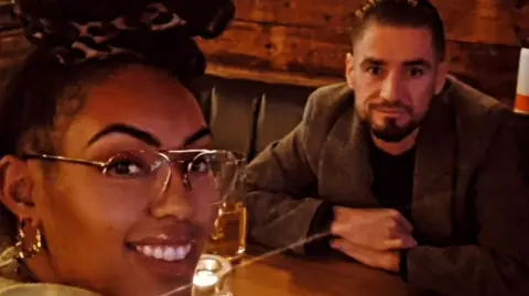 Sian is closet to the camera and smiles. She is wearing clear glasses and gold earrings. She is sat at a table opposite a man wearing a dark blazer and dark hair