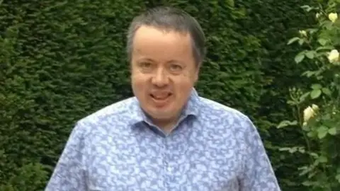 Family photo A man with grey hair smiling at the camera in front of green bushes. He is wearing a short sleeved blue patterned shirt.