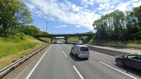 Two bridges can be seen over a dual carriageway. Trees and greenery can be seen on either side.