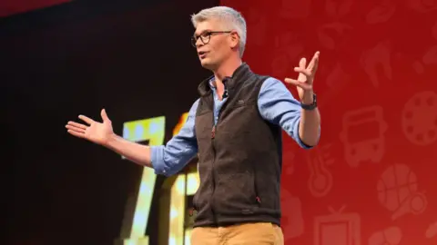 Getty Images MP Al Pinkerton stood on a stage with a red background. He is wearing a blue shirt and a brown sleeveless jumper. He has his hands aloft. 