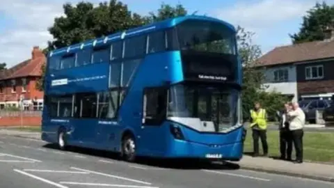 LDRS A blue electric bus is parked alongside a residential road, with three people standing next to it on the pavement