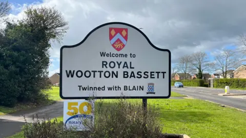 A sign which says 'Welcome to Royal Wootton Bassett' with a road behind it