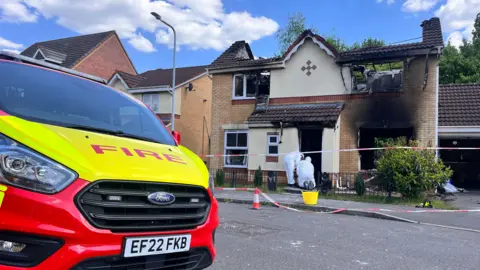 Forensic officers wearing white suits are outside a house that has been destroyed in a fire. It is a burnt-out shell with views through to the back garden. The forensic officers are working where the front door would have been. Red and white fire tape is outside. In the foreground there is a large vehicle with the word "FIRE" in red on a yellow bonnet.
