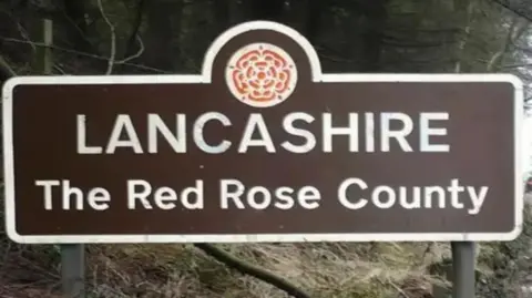 Large brown road sign with white writing. It reads "LANCASHIRE - The Red Rose County". The county's Red Red emblem is included.