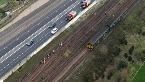 A drone shot showing a the train with two carriages stationary on the tracks. There are people walking on the tracks, some are wearing hi-vis clothing. There is a dual carriageway running along the tracks and there are two fire trucks and a white car parked while other cars are driving past.