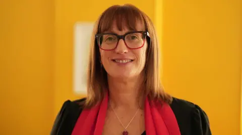 Julieanne Mitchell has reddish brown straight hair that falls just below her shoulders and a fringe above her red and dark brown-framed large glasses. She is looking directly into the camera and smiling, while wearing a red scarf and a black jacket, with a necklace that has a red heart pendant. The room behind her is painted bright yellow.