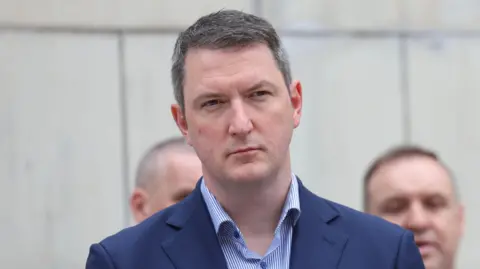 Pacemaker A still of John Finucane standing outside a white-stone building. He has short greying hair and is wearing a blue suit jacket as well as a blue and white striped shirt. The background is blurred.