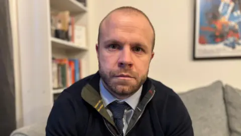 Hombre de la BBC con cabello corto y cabello facial con un jersey azul y camisa