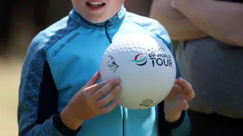 Getty Images A young fan, whose face is not visible, holds a giant autographed golf ball. He is wearing a light blue golf jacket with patterned sleeves. 
