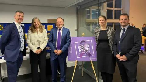 Five senior leaders stand in a line with an easel placed at the centre displaying the new strategy artwork - a purple board with white text reading “Gloucestershire Tackling Domestic Abuse Strategy 2025–2028.” From left to right stand: Nick Evans, Deputy Police and Crime Commissioner; Sarah Truelove, Chief Executive of NHS Gloucestershire Integrated Care Board; the easel; Cllr Paul Hodgkinson, Cabinet Member for Public Health; Siobhan Farmer, Director of Public Health; and Detective Superintendent Ian Fletcher, Gloucestershire Constabulary.