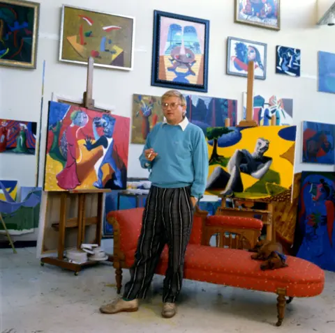 Getty Images Portrait of English painter David Hockney, dressed in a light blue sweatshirt and tan trousers, as he stands in front of a chaise longue in his home, Los Angeles, California, 1987.