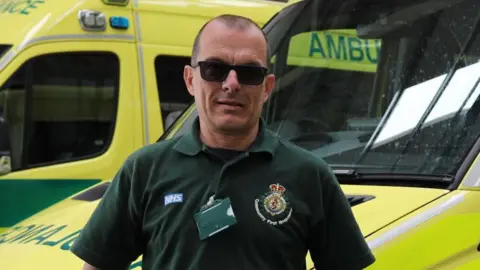 EEAST Stuart Little wearing a dark green polo top, with the Community First Responders' logo, and black sunglasses. He is standing in front of two ambulances.