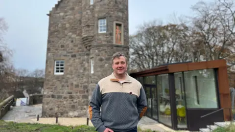 BBC A man is standing in front of the old brick tower with the modern looking extension building attached to it.