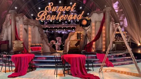 The set of BBC's Strictly Come Dancing studio set as the staging of the musical Sunset Boulevard. There are two round tables in the foreground with red cloths draped over them and falling onto the floor. To the right of the picture is an A frame set of wooden ladders 