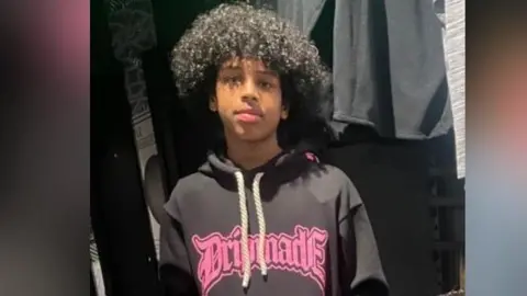 Family photo Mohanad Abdullaahi Goobe is photographed with black curly hair. He is pictured wearing a dark hoodie with bright pink writing on it.