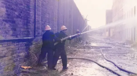 PA Media/handout Two men in dark overalls and yellow hard hats point a water hose towards a fire in what looks like an old industrial area.