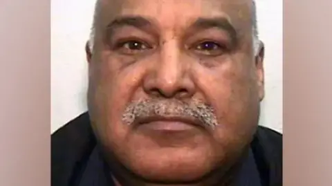 Police custody image of Shabir Ahmed who has a grey receding hair and moustache. He is wearing a black top. 
