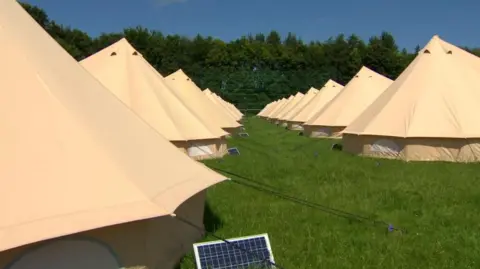 A number of tents are seen in a row in a field. A solar panel is visible from the side of the first tent.