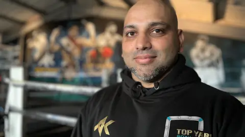 Emma Baugh/BBC Hamad wearing a black hoodie, looking at the camera or photographer. A boxing ring is behind him.