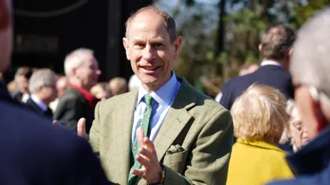 Zoe Head-Thomas/PA Wire The Duke of Edinburgh holding his hands out greeting someone by the camera. He is smiling, has short hair and is wearing a green/ brown blazer.