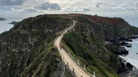 BBC A winding bridge with grassy outcrops sloping down on either side. Two cyclists are pushing their bikes on the bridge, with a tractor in the background. 