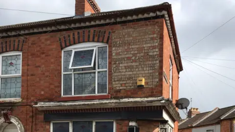 MOLA A two-storey red brick end of terrace house. It is on a street corner. Above the ground floor and beneath the roof is a very faded cream rectangle with words written on it, which are very hard to read.
