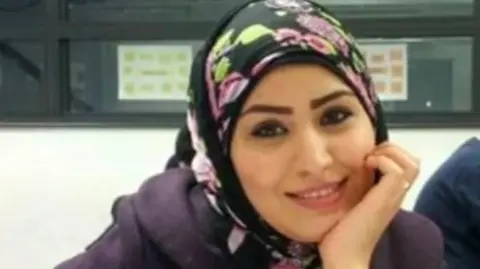 Rania Alayed smiling wearing a black hijab with pink flowers and purple suit jacket with her face resting on her hands