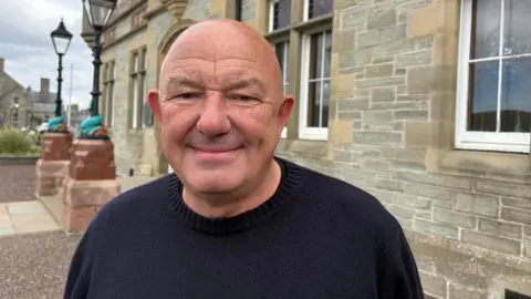 A bald headed man with a dark, round-neck jumper on stands in front of a granite building with two lamp posts on sandstone pillars behind him