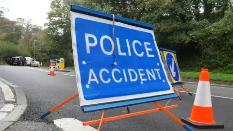 BBC A Police Accident sign is in the foreground next to a traffic cone while in the background there are vehicles on the road 
