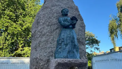 A statue of a woman on a large rock-style water fountain. There are tall trees in the background and the sky is clear blue.