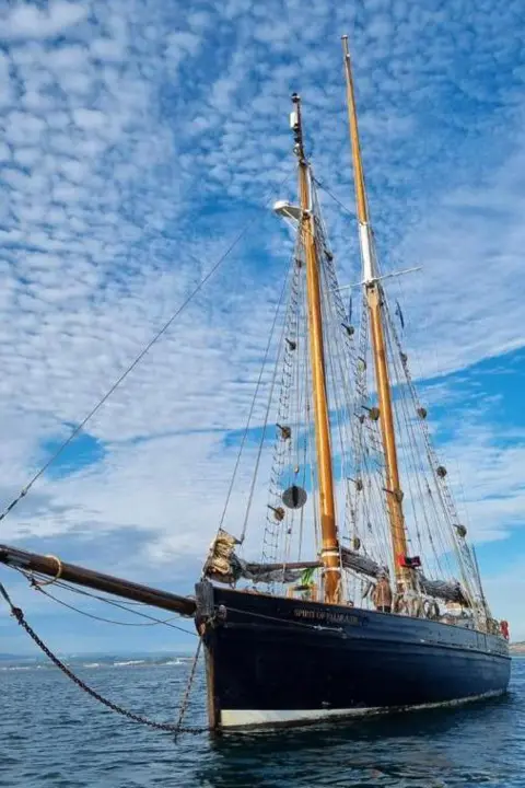 Turn to Starboard Spirit of Falmouth is a navy blue tall ship sailing with a mackerel sky above it and calm seas below. Her sails are furled.