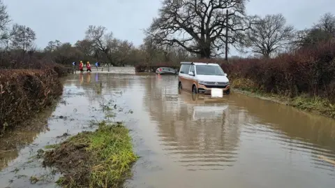 DWFRS Two vehicles stuck in floodwater - a gold and white camper van and a grey hatchback which is turned sideways and tipping forward. Firefighters in red uniforms and waders are walking in the distance.