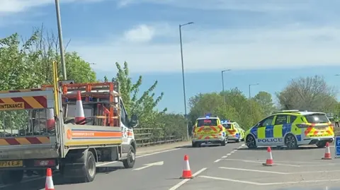 Part of the A10 where a road closure is in place. At the scene of the incident there are a number of marked police cars parked on the road and a highway maintenance van.