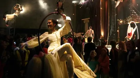 Colin Moody/ Plaster Aerial hoop performers in white suspended at various points in the air above and around a standing audience