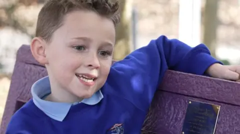 Teddy, is seen smiling as he sits on a purple bench made from recycled plastic tubs. He is wearing a light blue shirt under a blue top