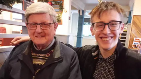 Walter Nightingale sits at a table in a restaurant next to his grandson Alexander who has his arm around him. Walter has thick, short white hair and wears glasses with black rims. He wears a patterned jumper under a grey coat. Alexander has short light hair, he wears glasses with black rims. Her wears a pattered shirt under a black jacket.