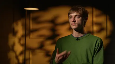Michael Gibson speaks during an interview, wearing a green jumper, with warm amber lighting and blurred shapes behind him.