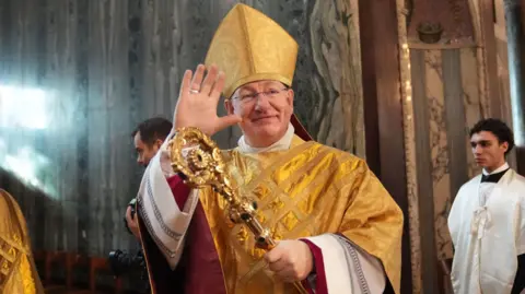 PA Media Bishop Richard Moth during his installation Mass as the 12th Archbishop of Westminster. He is smiling as he waves at onlookers. He is dressed in a gold robe and carries a gold crozier (pastoral staff)