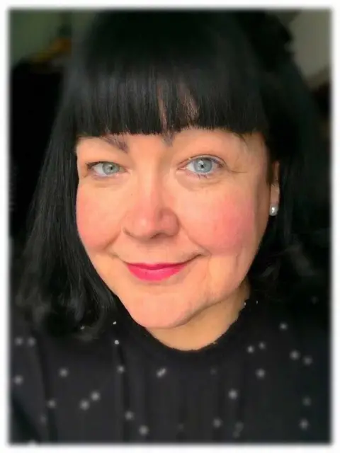 Campaign Against Living Miserably A woman with black hair and pearl earrings is looking into the camera. She is wearing a black blouse with white dots. 