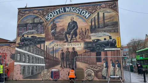 BBC A colourful mural featuring terraced houses, steam trains and Orson Wright with the words South Wigston at the top