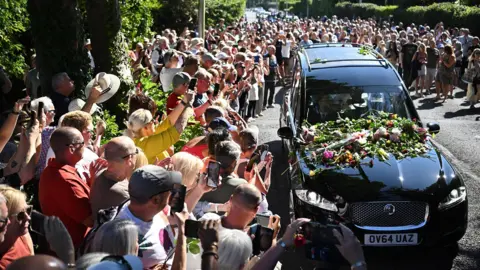 Getty Images Bystanders flock towards a black hearse and throw flowers onto its bonnet as it drives through the town carrying the coffin of Bonnie Tyler.