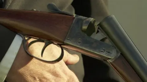 BBC A close-up of a classic double-barrel shotgun with break-action being held by a man wearing a black suit with a white shirt cuff visible. The firearm has a wooden stock, metal components and features two triggers. The shotgun is open, revealing the breech, and appears to be unloaded. 