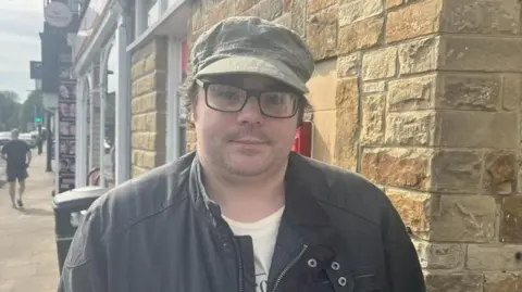 BBC/Rachel Russell A man wearing a dark coat, light t-shirt and green peaked hat stands at the corner of a stone building. He is wearing black-rimmed glasses and looking into the camera. A street can be seen stretching into the distance behind him.