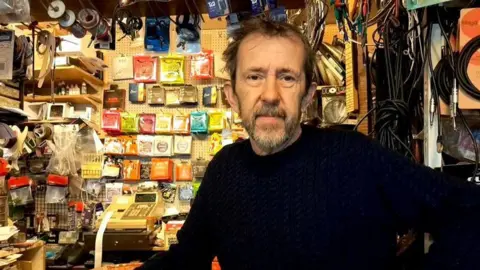 Gary Good at his store The Music Box on Cowley Road. He is looking at the camera. There are many cables and other music instruments behind him.